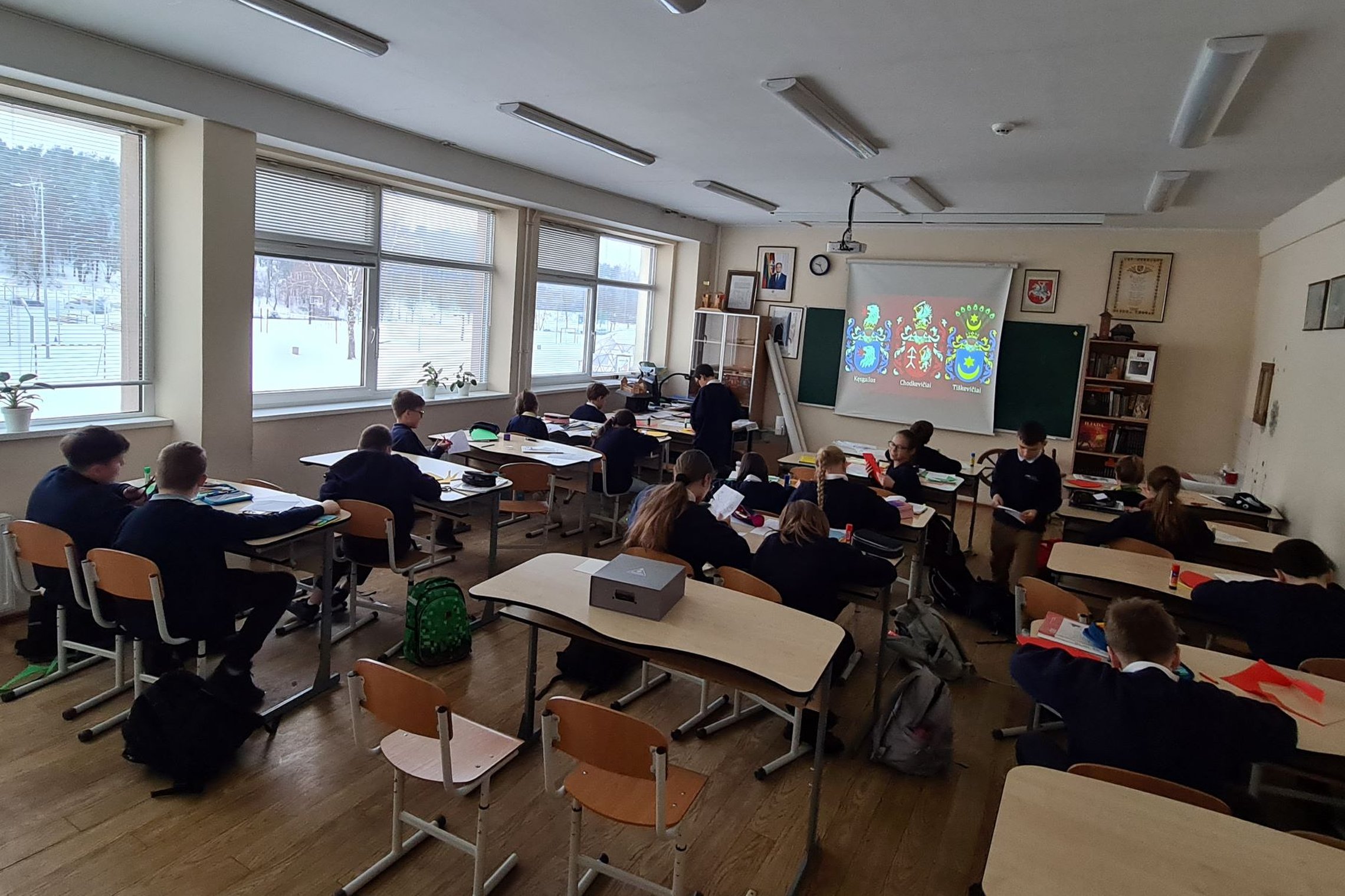 Vaikai klasėje fotografuoti iš nugarų. Projektoriuje matosi puošnūs herbai.