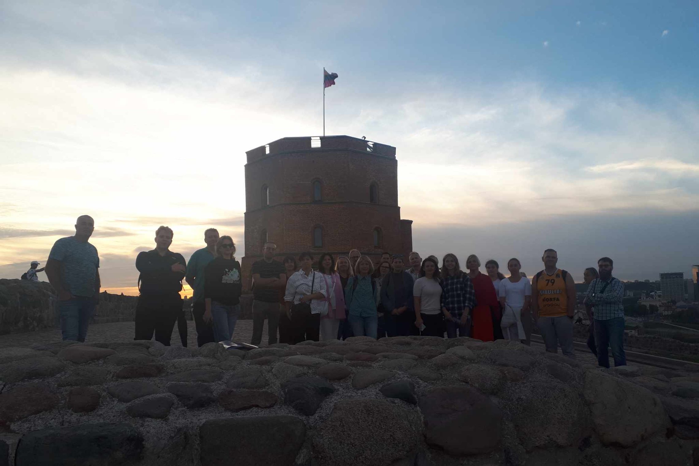 Ant Gedimino kalno stovi didelė grupė žmonių, už jų matome Gedimino pilies bokštą ir besileidžiančią saulę. Žmonės kiek atitolę nuo fotografo, prieš juos matosi žema akmenų siena.