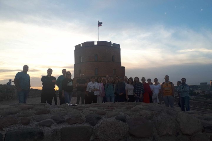 Ant Gedimino kalno stovi didelė grupė žmonių, už jų matome Gedimino pilies bokštą ir besileidžiančią saulę. Žmonės kiek atitolę nuo fotografo, prieš juos matosi žema akmenų siena.