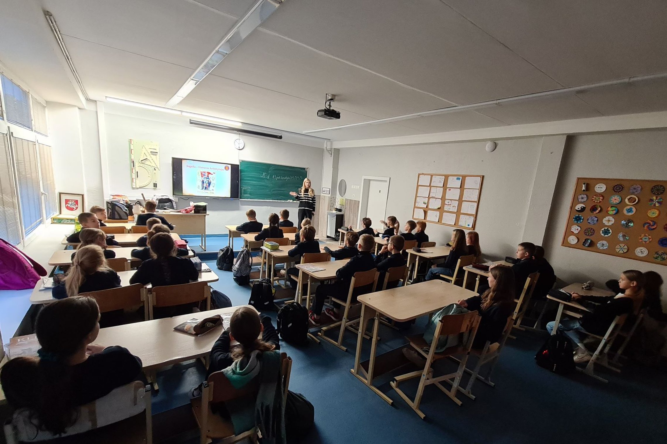 Klasė mokinių, sėdinčių suoluose. Klasėje prieblanda, vaikai žiūri į projektorių.
