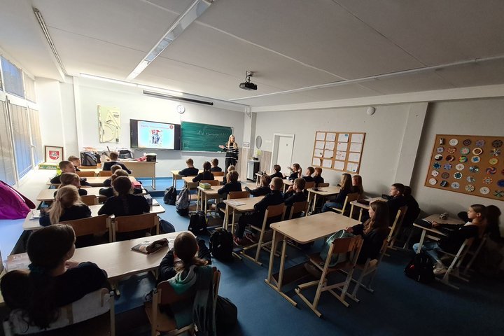 Klasė mokinių, sėdinčių suoluose. Klasėje prieblanda, vaikai žiūri į projektorių.