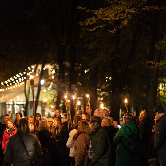 Grupė ekskursijos dalyvių eina per Pilių parką. Jų fone matosi Rotondos kavinės šviesos.