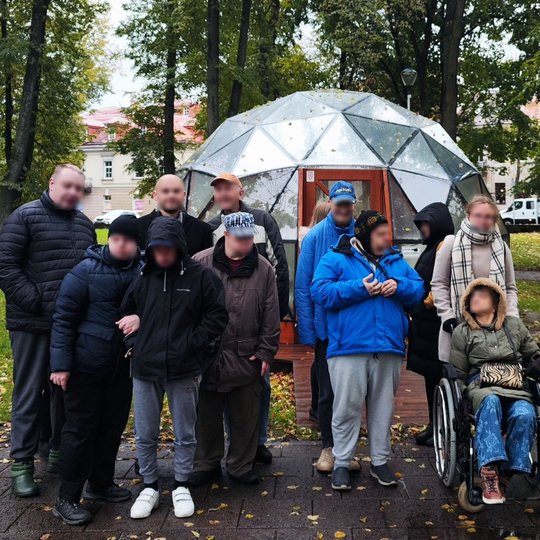 Žmonių grupelė stovi prie Vilniaus pilių kupolo, kuris yra Pilių parke. Už jų matosi keli miesto pastatai. Lauke  šlapia, lyja.