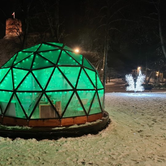 Vilniaus pilių kupolas iš lauko vakare. Kupolas šviečia žalia spalva. Kairėje, viršuje matosi Gedimino bokštas su plevėsuojančia trispalve.