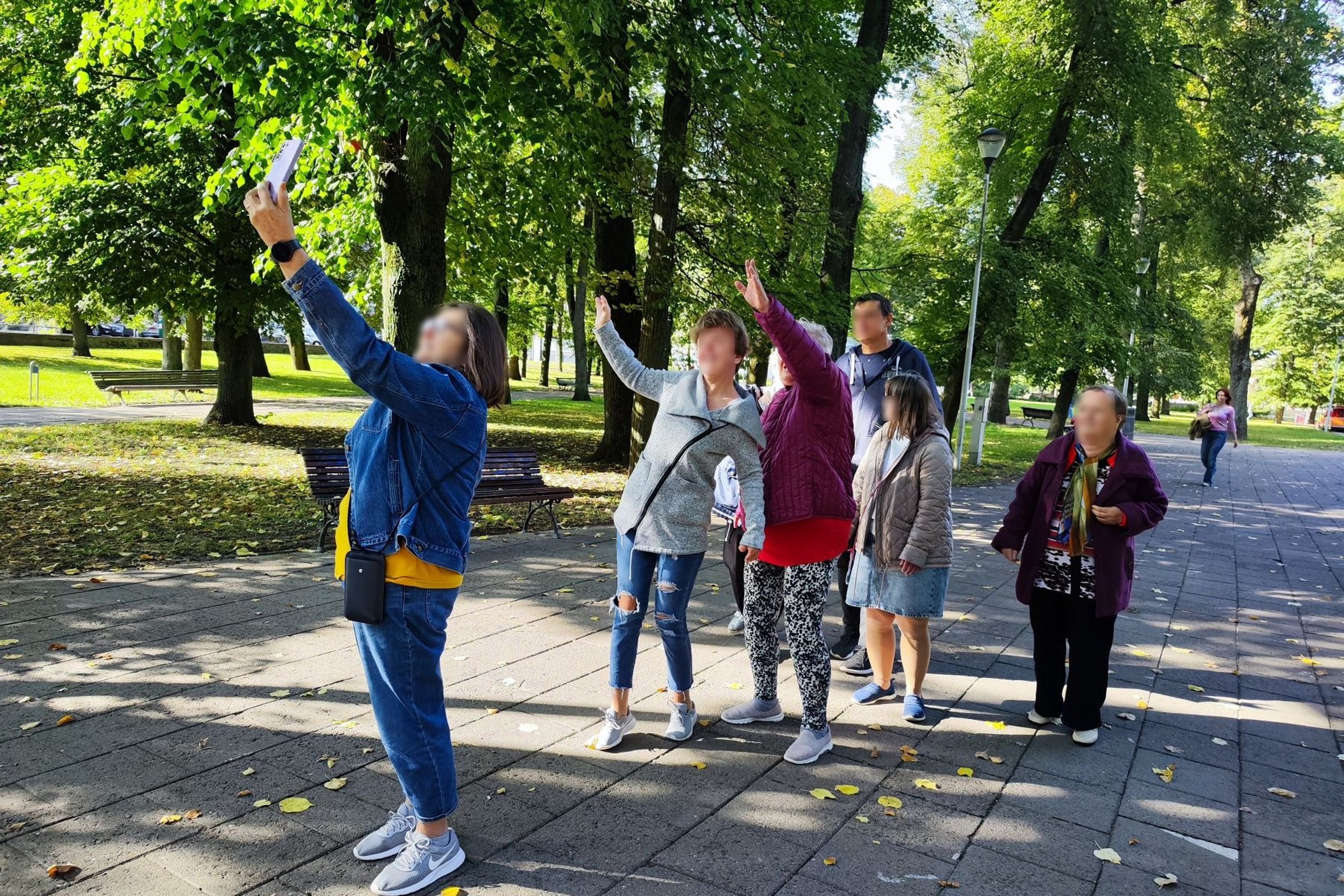 Nuotraukoje matome kelis žmones, kurie fotografuojasi parke, netoli Katedros aikštės. Vienas iš jų laiko telefoną, o kiti stovi jam už nugaros ir pozuoja.
