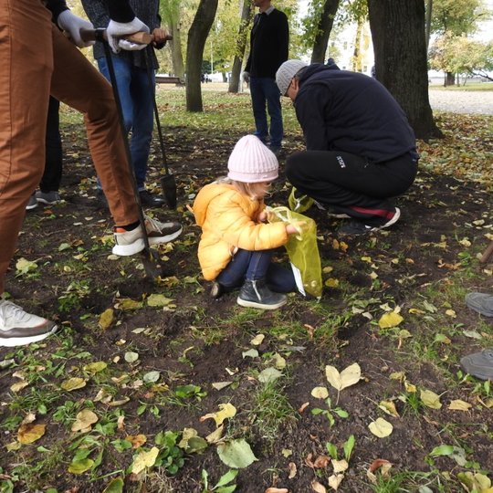 Maža mergaitė padeda sodinti gėlytės. Aplink dirba suaugusieji. 