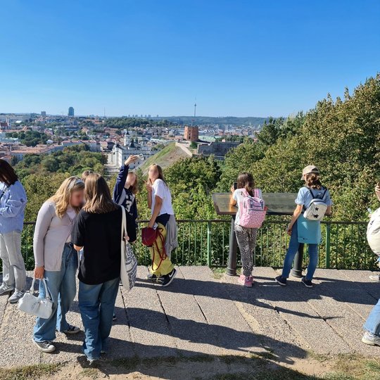 Giedra diena. Viduryje nuotraukos matome dvi besikalbančias mergaites, už jų dvi kitos mergaitės darosi asmenukę, iš šonų esantys žmonės mėgaujasi vaizdu nuo Trijų kryžių kalno.