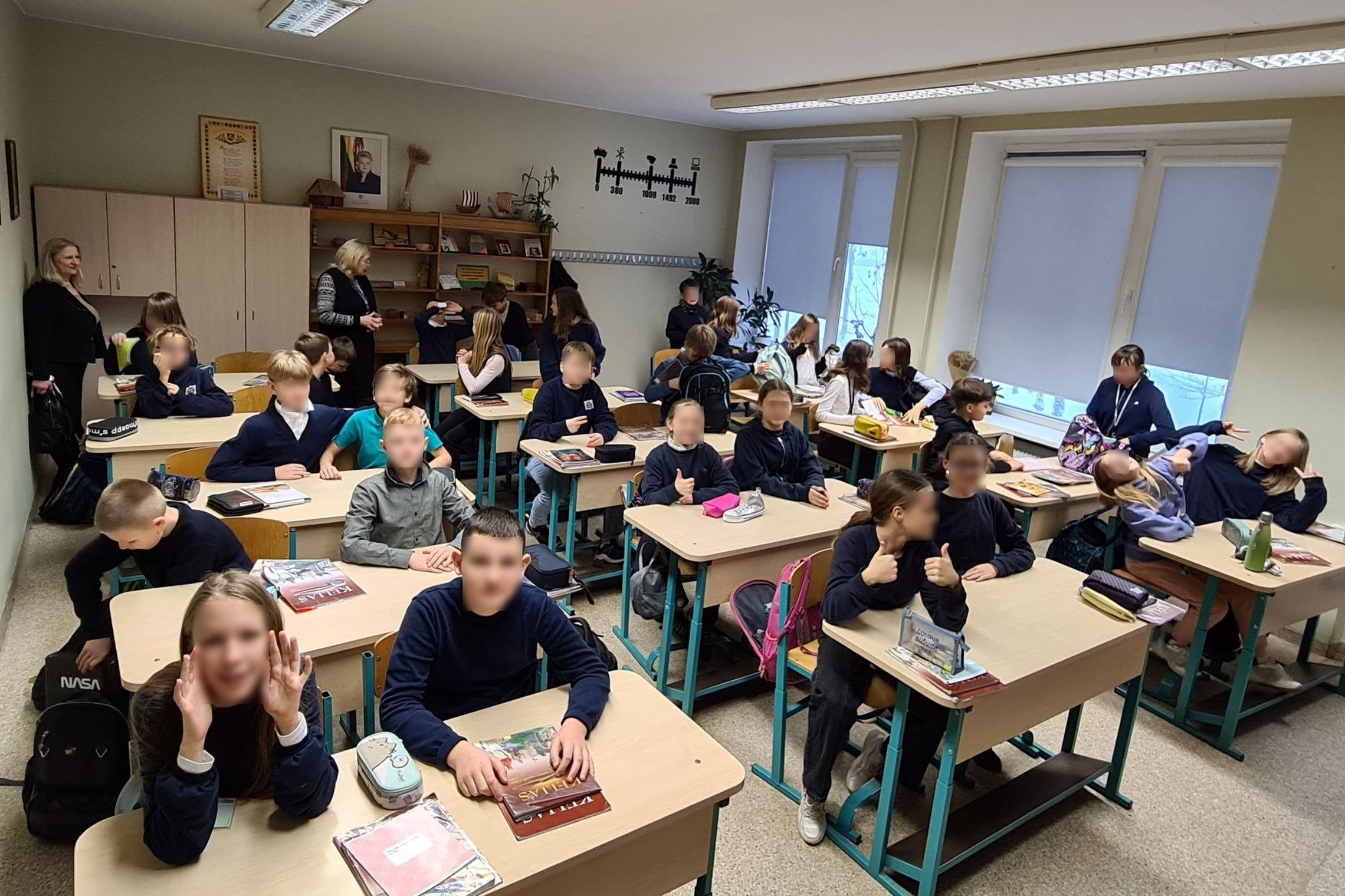 Penktokai sėdi klasės suoluose ir žiūri į objektyvą. Klasės gale stovi dvi mokytojos.