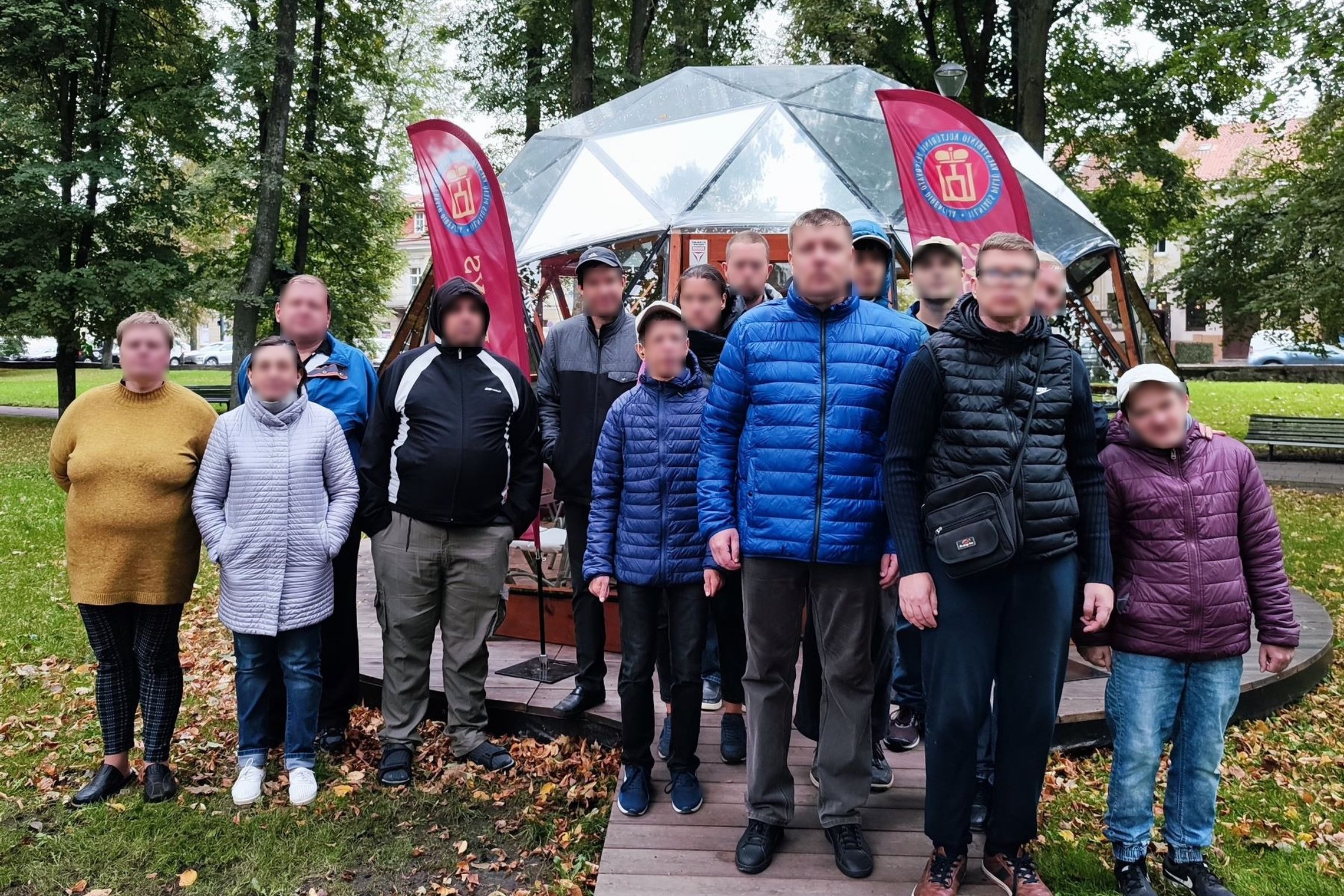Grupelė įvairaus amžiaus žmonių yra sustoję nusifotografuoti, tiesiai už jų matosi Vilniaus pilių kupolas.