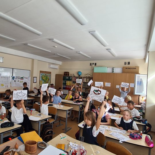 Nuotraukoje yra klasėje sėdintys trečiokai, iškėlę į orą savo piešinius. Jų stalai nukrauti įvairiomis priemonėmis, ant sienos kabo trys mokyklinės lentos su plakatais, klasės gale stovi spinta su knygomis.