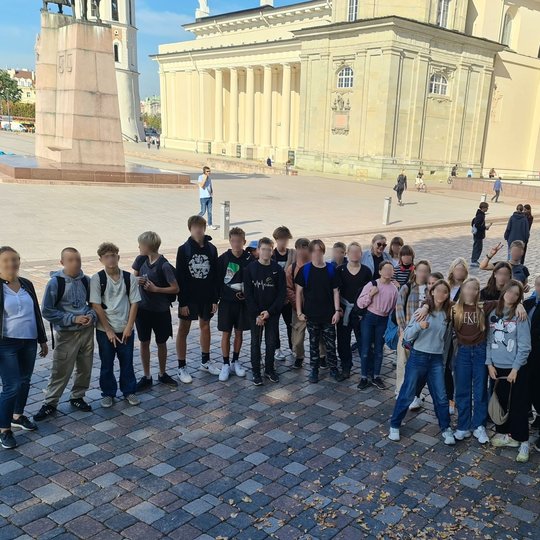 Nuotraukoje 30 septintokų, kurie yra sustoję bendrai nuotraukai Katedros aikštėje. Visi šypsosi. Katedros aikštėje labai saulėta, tačiau jų fotografijai netrukdo saulė, nes visi atsistojo pavėsyje.