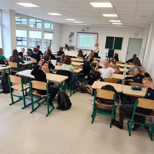 Nuotrauka daryta iš klasės galo. Klasėje 30 penktokų, kuriems mokytoja aiškina svarbią temą, tačiau paskutinė suolų eilė atsisukus į kamerą, nesiklauso mokytojos, jie iškėlę du pirštukus - V formos.