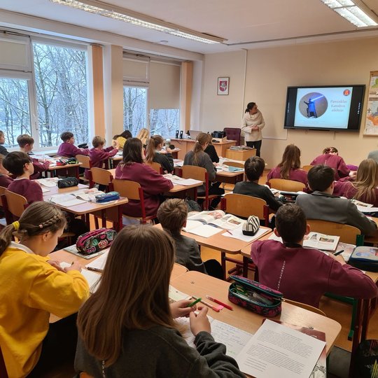 Klasėje, pilnoje mokinių, vyksta istorijos pamoka. Priekyje klasės stovi edukatorė.