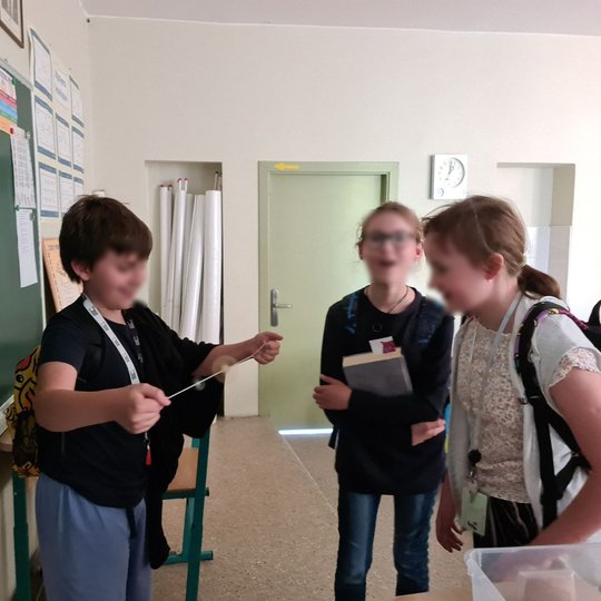 Nuotraukoje vaizduojami klasėje stovintys trys vaikai. Berniukas žaidžia su besisukančia saga ant virvutės, viena mergaitė stebi, abu šypsosi. Trečioji mergaitė žiūri į kamerą su šypsena. Vaikai stovi prie mokyklinės lentos su užrašu “Archeologija - kas t