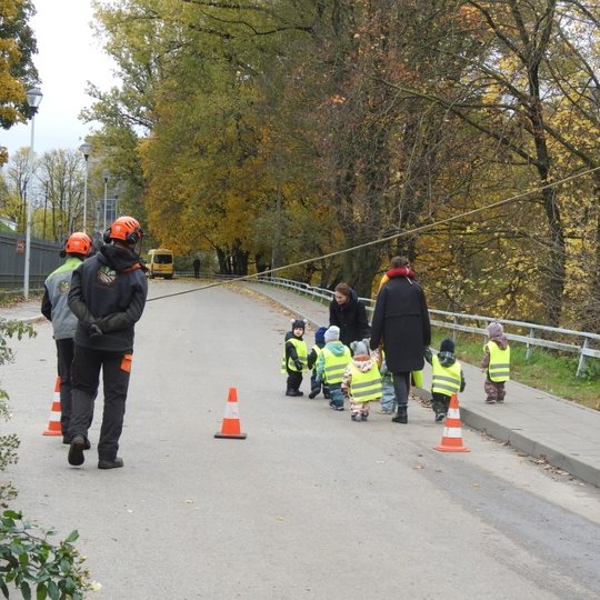 Ant kelio, apstatyto eismo kūgiais, vaikščioja maži vaikai su atšvaitinėmis liemenėmis, lydimi dviejų auklių. Paskui šį būrį eina darbininkai.