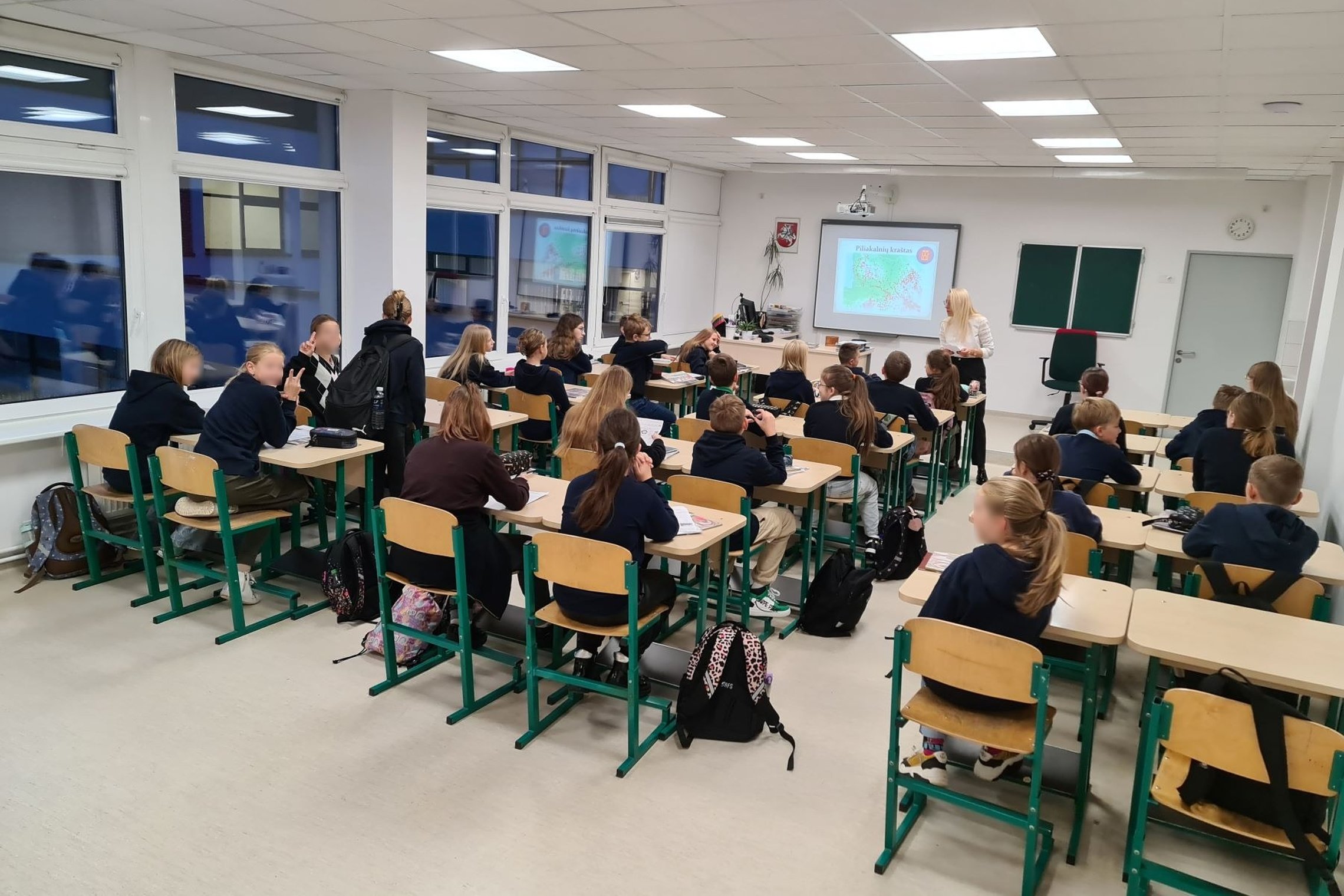 Nuotraukoje matome progimnazijos mokinius, kurie įdėmiai žiūri ir klausosi mokytojos pasakojimo. Nuotrauka padaryta iš klasės galo. mokytoja yra įjungusi skaidres ir rodo prezentaciją. Kai kurie vaikai pozuoja kamerai.