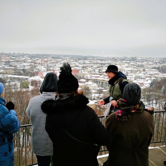 „Lietuvos nacionalinis kultūros centras“ kolektyvas priešais Trijų Kryžių paminklą klausosi gido pasakojimo. Už nugaros matosi Vilniaus panorama su apsnigusiais stogais.
