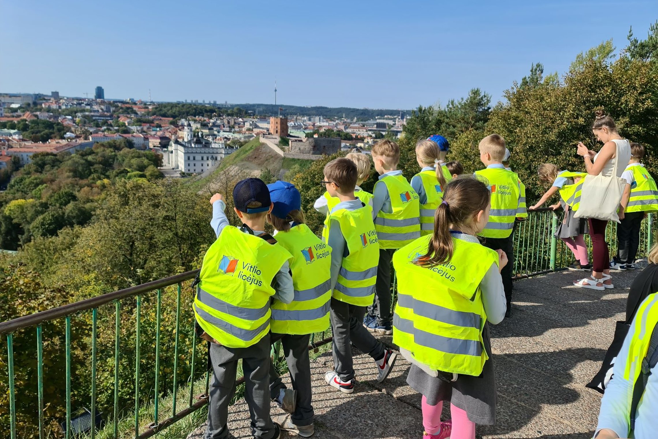 Aukštai ant kalvos stovintys antros klasės mokiniai, dėvintys ryškiai geltonas liemenes su užrašu "Vitlio licėjus", stebi Vilniaus miesto panoramą, kurioje matoma Gedimino pilis, Katedros aikštė, senamiestis.