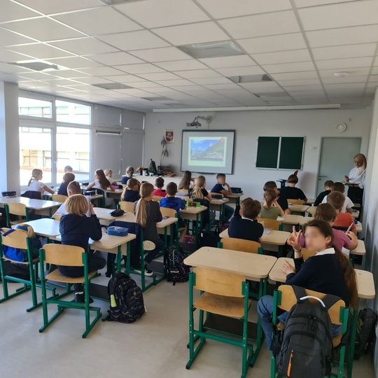 Nuotraukoje matome penktos klasės moksleivius. Mokiniai stebi mokytojos pateiktas skaidres. Mokytoja stovi arčiau durų.