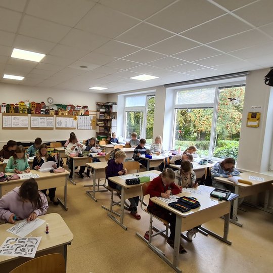 Nuotraukoje vaizduojama klasė mokinių. Kiekvienas mokinys ant suolo turi po du lapus. Visi mokiniai susikaupę dirba, vieni kažką klijuoja lapuose, kiti kerpa, o dar kiti piešia.