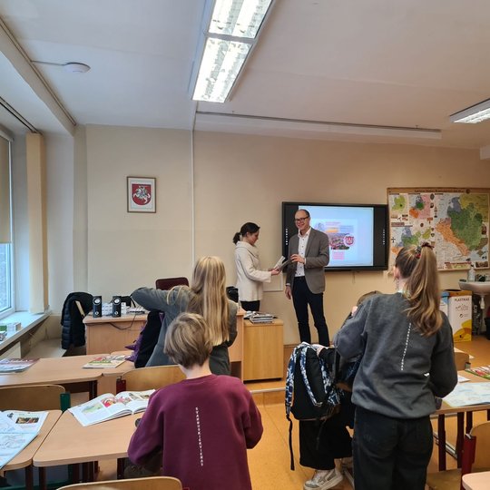 Mokytojas su edukatore veda istorijos pamoką. Vaikai sėdi ir stovi prie suolų.