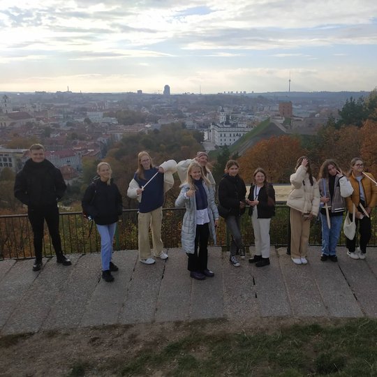 Ekskursijos dalyviai pozuoja nuotraukai nuo Trijų Kryžių paminklo. Jiems už nugaros matosi Gedimino kalnas su bokštu ir Vilniaus miesto panorama.