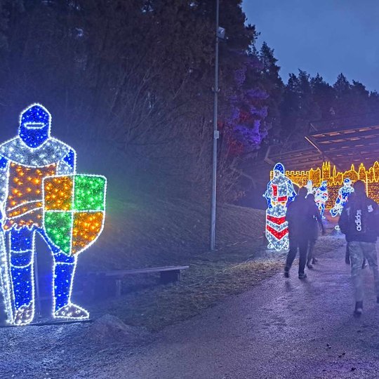 Grupė savanorių keliauja į Kreivosios pilies instaliaciją estradoje. Tako šonuose stovi šviečiantys kariai.