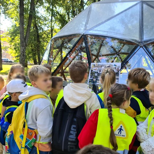 Nuotraukoje vaikų grupė su ryškiomis, geltonomis liemenėmis. Vaikai stovi parke, prie stiklinio kupolo, ant kurio prikabinta informacija, vaikai ją skaito.