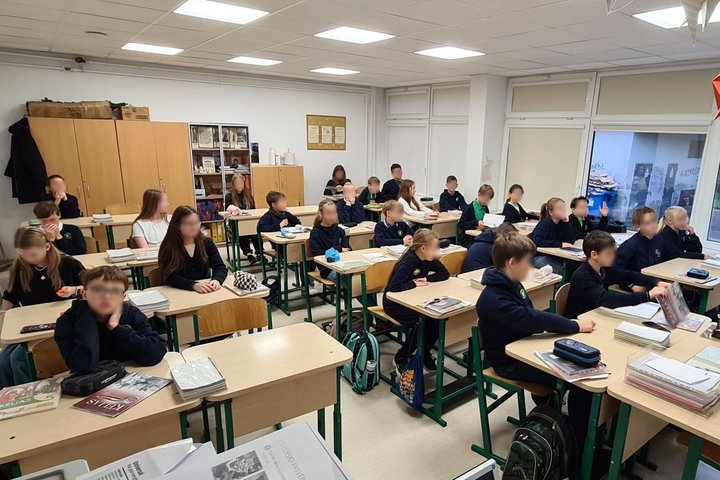 Nuotraukoje vaizduoja klasėje prie savo suolų sėdinčius penktokus. Nei vienas iš jų nežiūri tiesiai į kamerą, bet klausosi savo mokytojos, kurios nuotraukoje nesimato. 