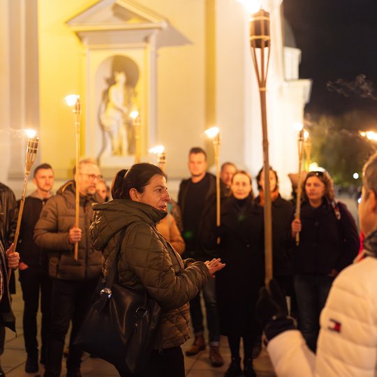 Gidė tarp ekskursijos dalyvių, kurie laiko fakelus.