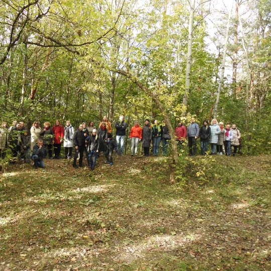 Žmonių grupė pozuoja nuotraukai miškelyje. Priešais juos yra pakankamai nemaža pievelė. Visi labai šiltai apsirengę ir sustoję vorele. Saulė silpnai šviečia, už jų puslankiu fone matosi medžiai.