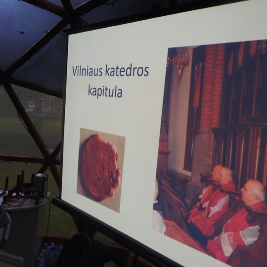 Vilniaus pilių kupole rodoma prezentacija. Skaidrėje sudėtos dvi nuotraukos, vienoje iš jų - vaškinis antspaudas su įspaustu kapitulos simboliu. Antroje nuotraukoje - trys kapitulos nariai sėdi bažnyčioje. Kairėje pusėje virš antspaudo yra užrašas “Vilnia