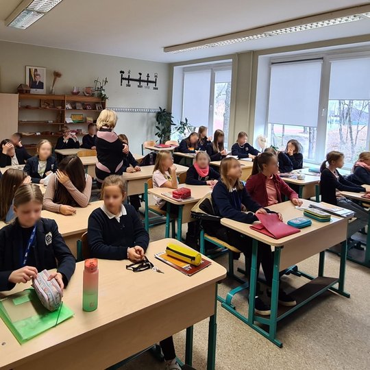 Penktokai sėdi klasės suoluose. Tarp jų stovi mokytoja.