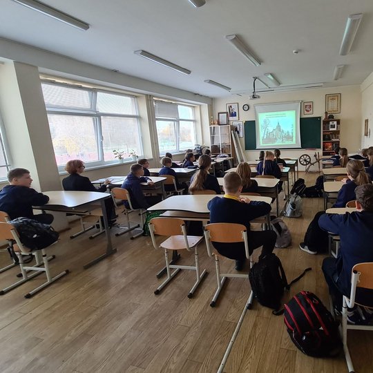Klasė, nufotografuota iš galo. Joje vaikai klausosi pamokos.
