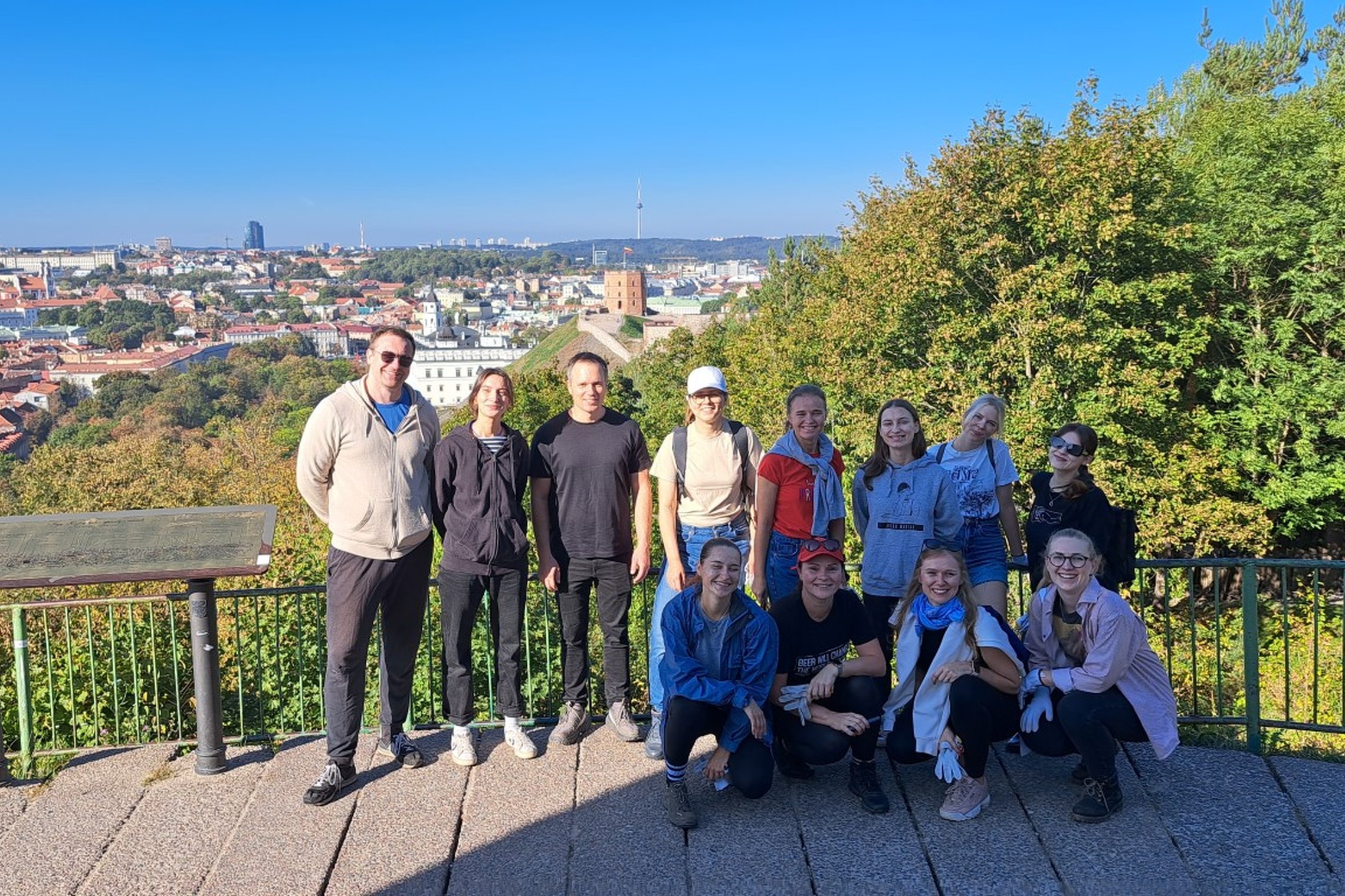Žmonių grupelė stovi ant Trijų kryžių kalno, pozuoja, už jų stūkso graži Vilniaus senamiesčio panorama. Žmonės šypsosi sustoję eilute, šalia vienas kito.