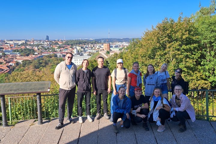 Žmonių grupelė stovi ant Trijų kryžių kalno, pozuoja, už jų stūkso graži Vilniaus senamiesčio panorama. Žmonės šypsosi sustoję eilute, šalia vienas kito.