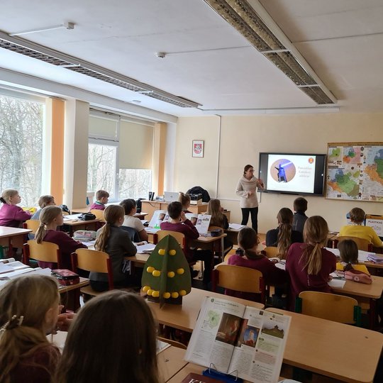 Edukatorė klasėje, pilnoje vaikų, veda istorijos pamoką.