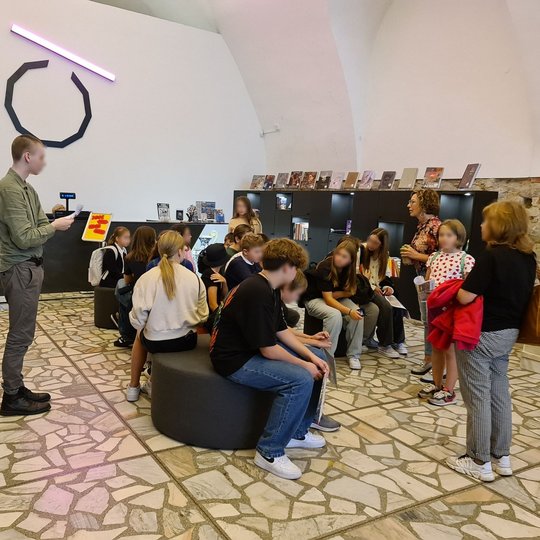 Nuotraukoje vaizduojami mokiniai muziejuje. Didžioji dalis sėdi, keli stovi.