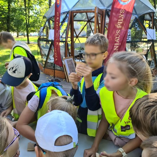 Nuotraukoje yra vaikai prie kupolo. Jie visi yra susibūrę prie stalo. Aplinkoje matosi ryškiai žalia žolė, daug medžių ir takelai.