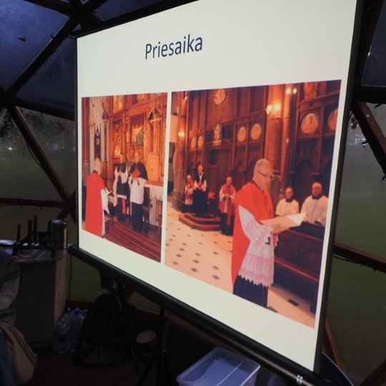 Vilniaus pilių kupole rodoma prezentacija. Šalia ekrano sėdi viena iš dalyvių, su susidomėjimu žiūrinti į ekraną. Skaidrėje įdėtos dvi nuotraukos, virš jų parašytas žodis “priesaika”. Nuotraukose vaizduojamos apeigos bažnyčioje.