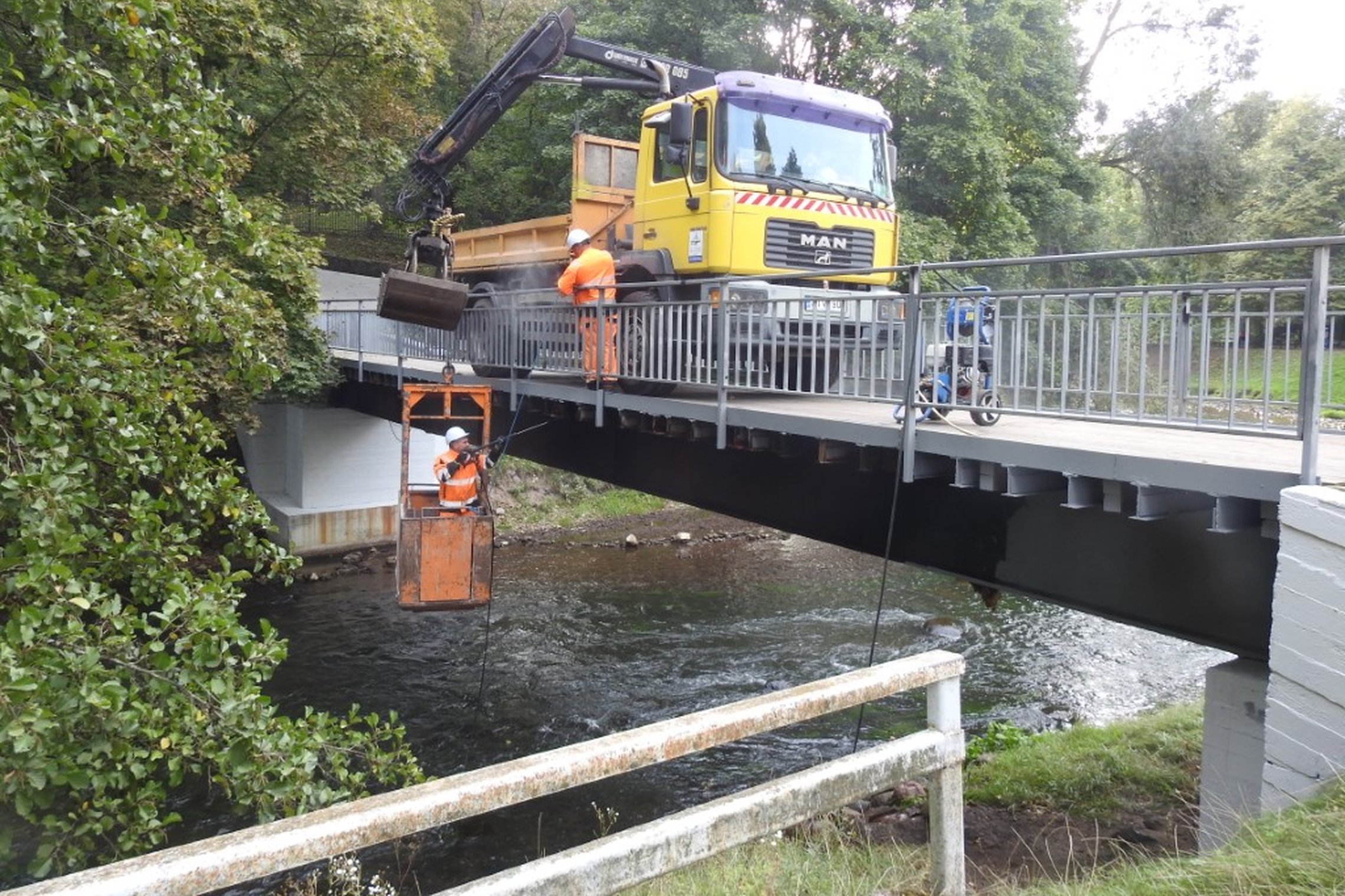 Vaizduojamas nedidelis tiltas per Vilnios upę. Ant tilto dirba du darbininkai su apsaugos šalmais. Taip pat šalia jų stovi vieno kaušo geltona sunkioji savaeigė mašina. Šis kaušas nuleistas kartu su vienu iš darbininkų, kad jis nudažytų tilto apačią - tai