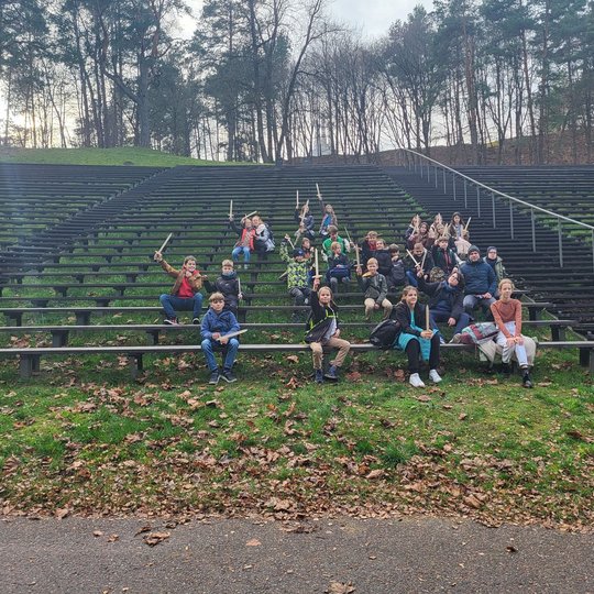 Penktokų klasė pozuoja nuotraukai sėdėdami ant medinių suolų Dainų slėnio estradoje.