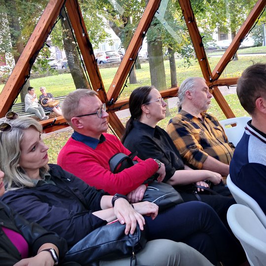 Kupole matome sėdinčių žmonių grupelę, kuri įdėmiai žiūri į prezentaciją, esančią už kadro.