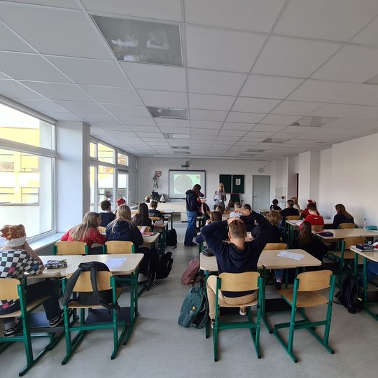 Mokiniai klasės suoluose, fotografuoti iš galo. Priekyje klasės stovi mokytoja.