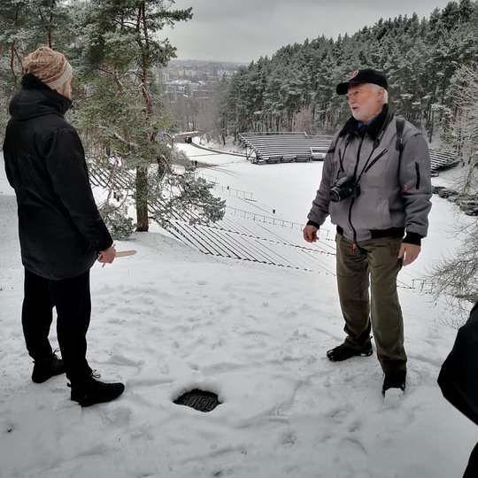 Edukatorius Stasys ant Karigailos kalvos intensyviai pasakoja ekskursantui apie Kreivąją pilį. Toliau matosi apsnigta Dainų Slėnio estrada.
