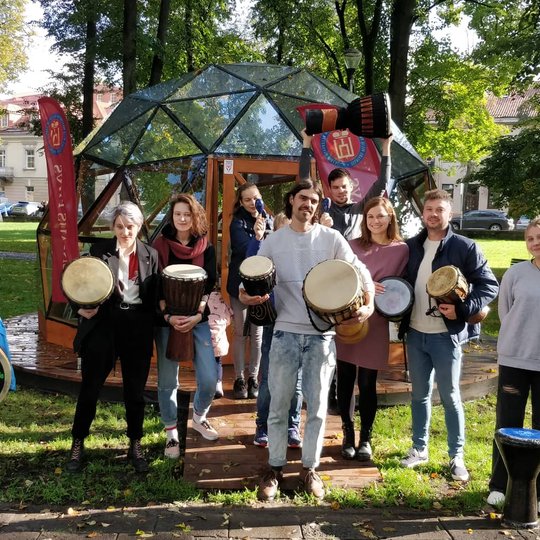 Būgnų rato dalyviai pozuoja nuotraukai su būgnais rankose priešais Vilniaus pilių Kupolą.