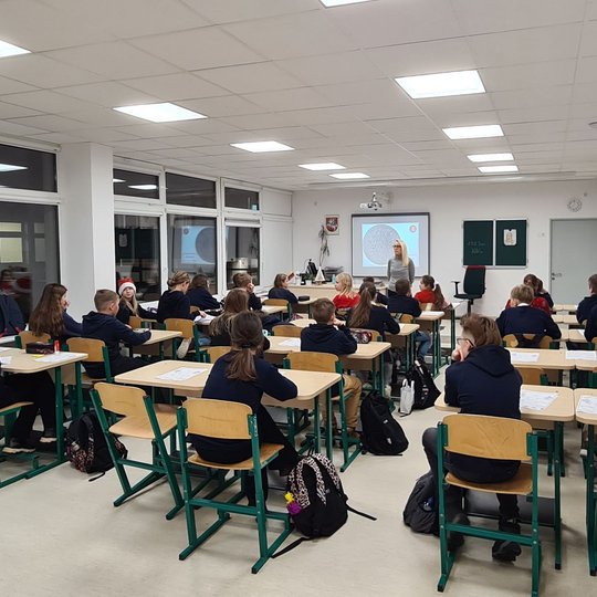 Mokiniai klasės suoluose, fotografuoti iš galo. Priekyje klasės stovi mokytoja.