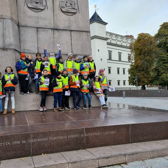 Ekskursijoje dalyvavę mokiniai džiaugsmingai pozuoja prie Gedimino paminklo Katedros aikštėje.