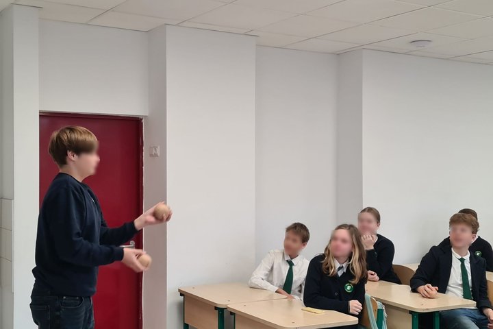 Nuotrauka vaizduoja klasę mokinių, kurie sėdi prie savo suolų, vienas iš jų stovi priešais savo klasiokus, kalba.