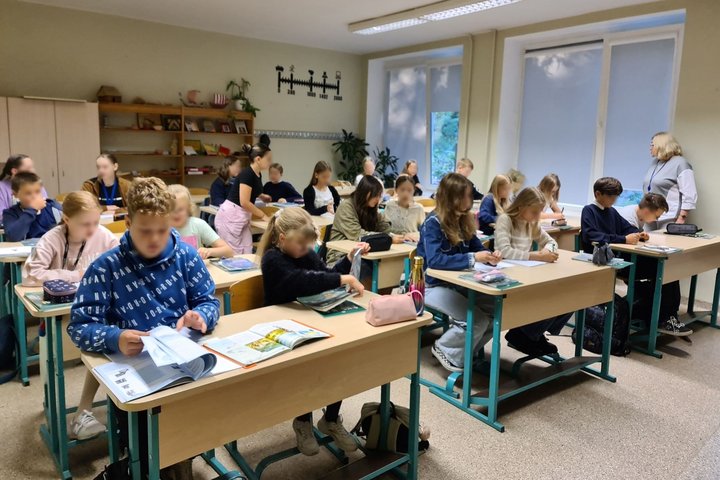 Nuotraukoje matomi mokyklos suoluose sėdintys mokiniai, kurie sėdi atsivertę vadovėlius ir mokosi. Galima pastebėti, kad mokytoja jiems dėsto temą. 
